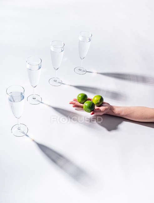 Female hand holding green ripe limes near clear glasses of water on white background — Stock Photo