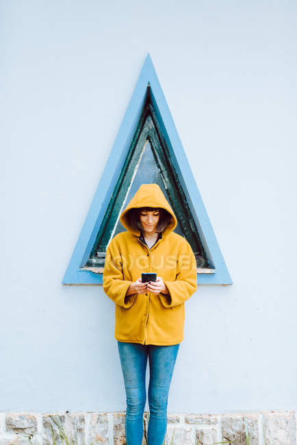 Jeune femme en manteau jaune chaud souriant et regardant vers le bas tout en se tenant contre la fenêtre triangulaire et le mur gris du bâtiment — Photo de stock