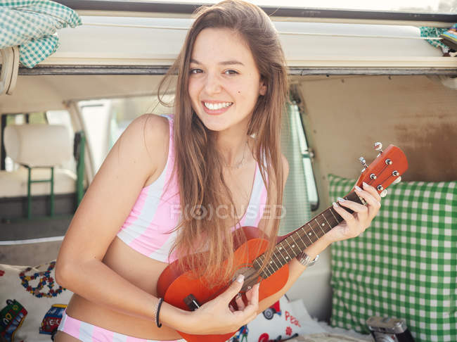 Mujer sonriente en traje de baño disfrutando jugando ukelele sentado en almohada suave en el coche en el fondo, mirando a la cámara - foto de stock