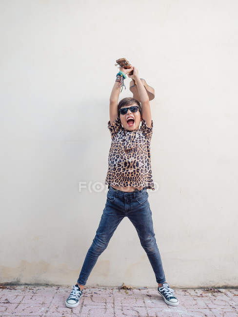 Niedlich fröhlich energisch fröhlich fröhlich Kind in Jeans und festlichem T-Shirt Spaß beim Spielen Ukulele auf dem Hintergrund der weißen Wand — Stockfoto