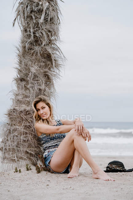 Portrait of young beautiful blonde seductive woman sitting down on beach and looking at camera — Stock Photo