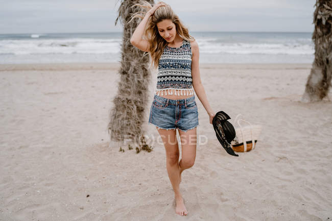 Attractive woman wearing top and shorts walking on sandy seashore with black hat — Stock Photo