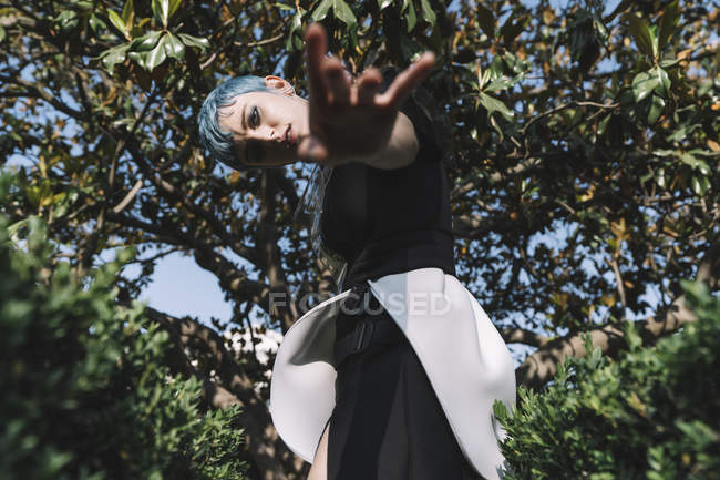 Von unten junge Frau mit kurzen blauen Haaren und in futuristischem Kleid, die sich nach vorne beugt und versucht, die Kamera auf verschwommenem Hintergrund zu berühren — Stockfoto