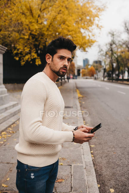 Giovane alla moda in piedi con il telefono cellulare sul ciglio della strada e la cattura di auto in autunno — Foto stock