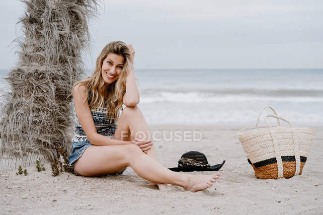 Porträt einer jungen schönen blonden verführerischen Frau, die sich an den Strand setzt und in die Kamera blickt — Stockfoto