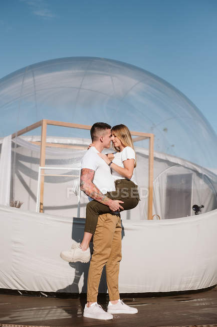 Vista lateral do homem apaixonado segurando e abraçando a jovem enquanto está na frente de glamping transparente romântico — Fotografia de Stock