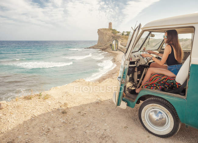 Vista lateral do carro com porta aberta e mulher desenhando na praia vazia arenosa com ondas espumosas em dia ensolarado brilhante — Fotografia de Stock