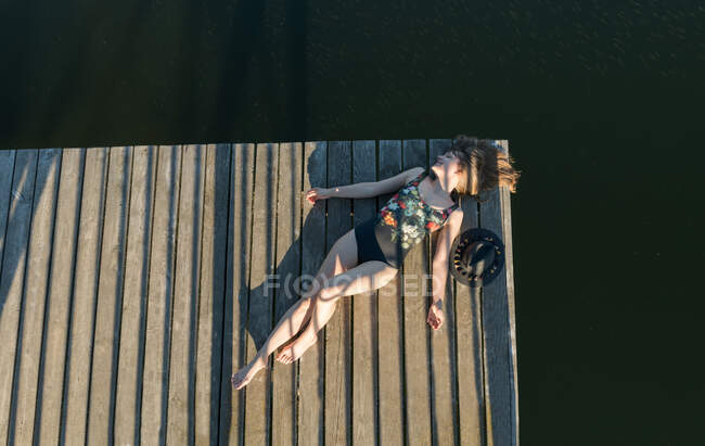Vista superior de la hermosa mujer en traje de baño negro y sombrero acostado en el muelle de madera del lago en el cielo azul claro y el fondo del bosque - foto de stock