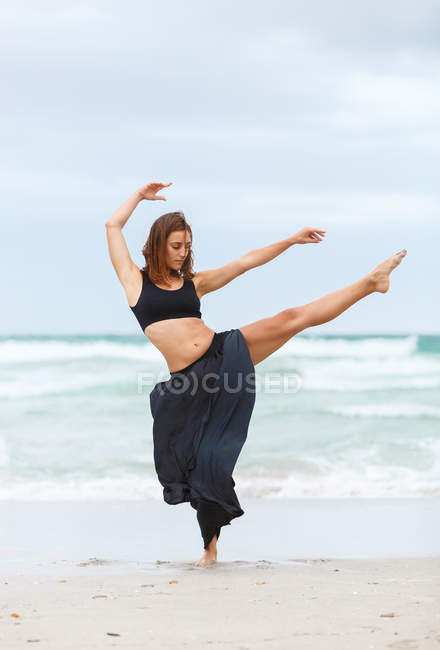 Fêmea atraente em roupa preta dançando na areia perto do mar ondulando — Fotografia de Stock