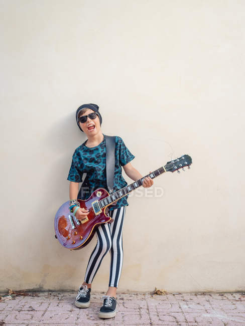 Atrevido ativo animado alegre menino em roupas coloridas tocando guitarra no fundo da parede branca — Fotografia de Stock
