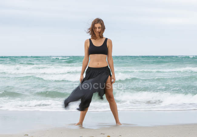 Confident young woman in black outfit standing on sand near waving sea — Stock Photo