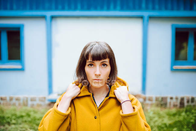 Jovem mulher de casaco quente amarelo sorrindo e olhando para a câmera enquanto está de pé contra a construção da parede cinza — Fotografia de Stock