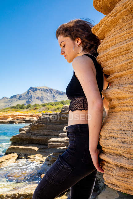 Mulher magra em top de cultura preta e jeans encostados contra a rocha na costa do mar — Fotografia de Stock
