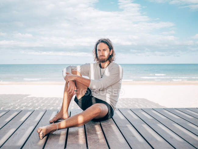 Uomo adulto barbuto seduto rilassato, appoggiato su un molo di legno in riva al mare guardando altrove — Foto stock