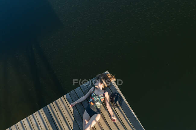 Vista superior da mulher bonita em maiô preto e chapéu deitado no cais de madeira do lago no céu azul claro e fundo da floresta — Fotografia de Stock