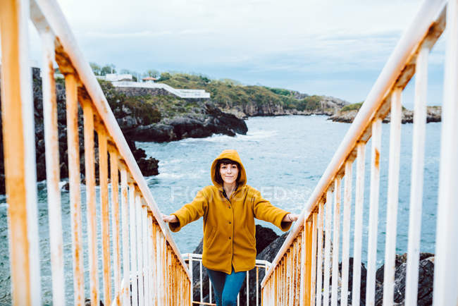 Молода жінка в жовтій куртці стоїть на іржавих сходах проти бухти з хвилястою морською водою — стокове фото
