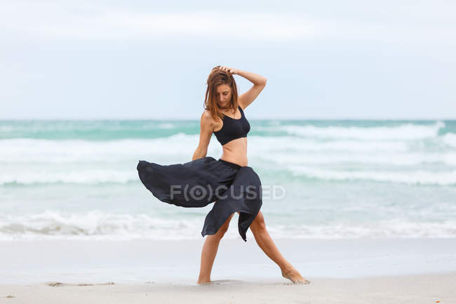 Привлекательная женщина в черном наряде, позирующая на песке возле моря — стоковое фото