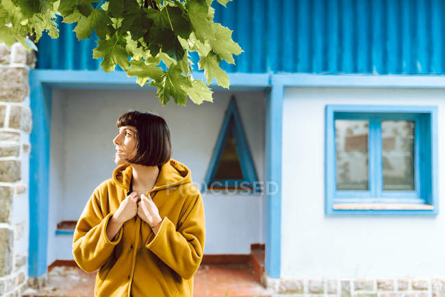 Junge Frau in gelbem warmen Mantel lächelt und schaut weg, während sie vor grauer Wand steht — Stockfoto