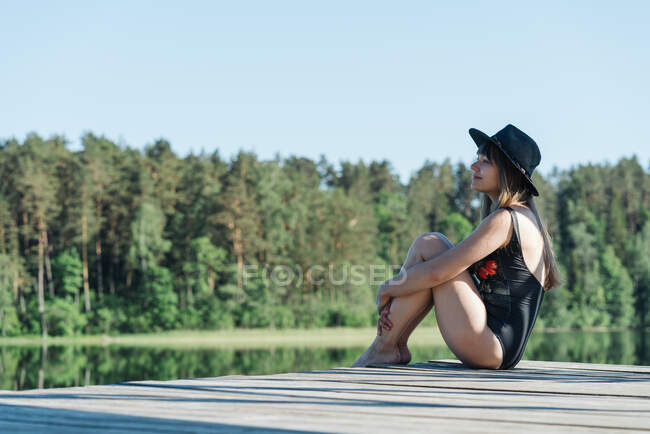 Vue latérale d'une jeune femme heureuse en maillot de bain noir et chapeau assis sur une jetée en bois et vue imprenable sur le lac sur un ciel bleu clair et un fond de forêt — Photo de stock