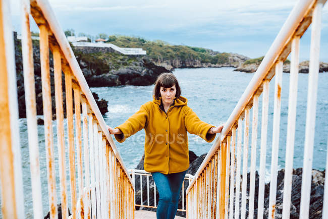 Jovem mulher de casaco amarelo em pé na escada enferrujada contra a baía com água do mar ondulando — Fotografia de Stock