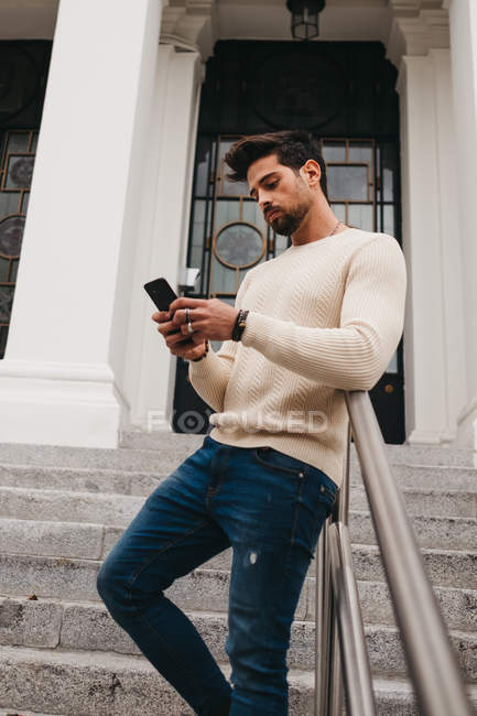 Homem barbudo elegante em pé nas escadas, inclinando-se sobre trilhos e usando telefone celular ao ar livre — Fotografia de Stock