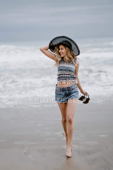 Mulher atraente em chapéu preto segurando saco de praia e sapatos enquanto desfruta de uma vista pitoresca do oceano olhando para longe — Fotografia de Stock