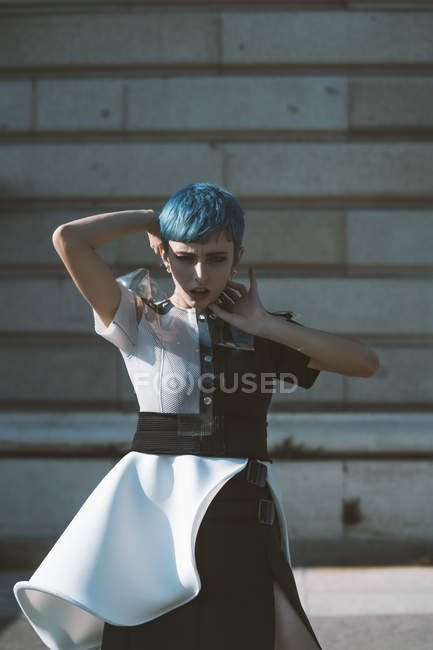 Young woman in futuristic dress touching face and short blue hair while standing near building on sunny day on city street — Stock Photo