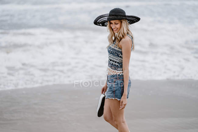 Attractive femme en chapeau noir tenant sac de plage et chaussures tout en profitant d'une vue pittoresque sur l'océan regardant vers le bas — Photo de stock