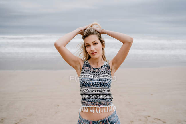 Fröhliche blonde Frau in buntem Top und Jeanshosen posiert, während sie sich am Strand entspannt — Stockfoto
