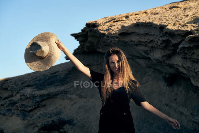 Young smiling woman looking at camera holding hat on rocky coast — Stock Photo