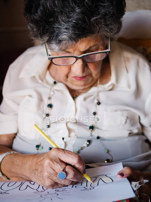 Пожилая седовласая женщина в белой рубашке и очках рисует на бумаге карандашом, сидит на кресле дома — стоковое фото