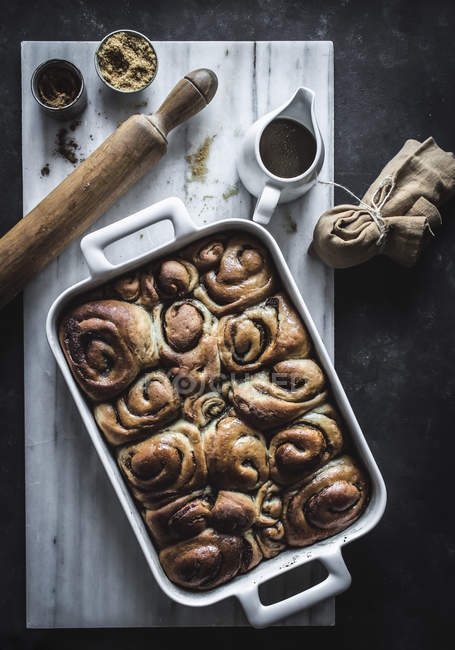 From above tray with fresh homemade cinnamon buns and wooden rolling pin on white marble board — Stock Photo