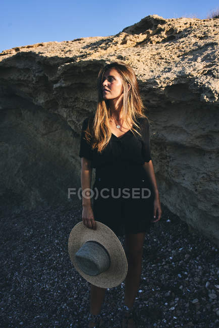 Young long haired smiling female model looking away holding hat on rocky coast — Stock Photo