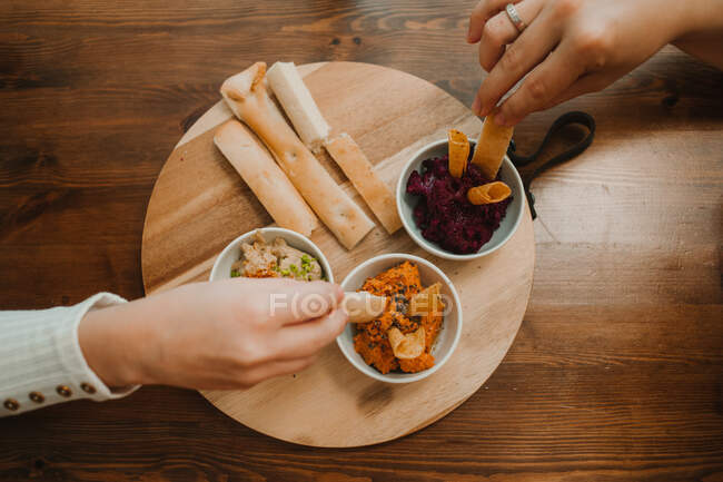 Из выше урожая неузнаваемые руки хватают разнообразные овощные соусы с хрустящим хлебом в миске на круглом столе — стоковое фото