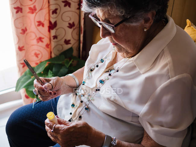 Mulher de cabelos grisalhos sênior em camisa e óculos segurando carretel de linha, sentado em poltrona em casa — Fotografia de Stock