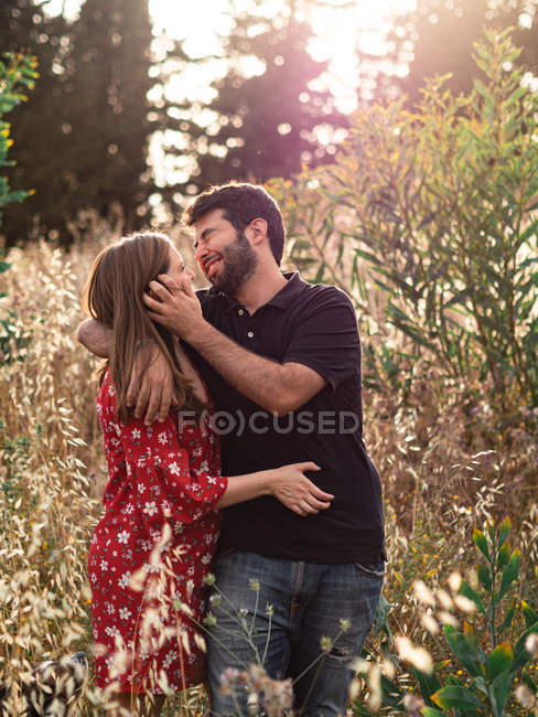 Заботливый мужчина обнимает улыбающуюся беременную жену на фоне живописного зеленого парка в солнечный день — стоковое фото