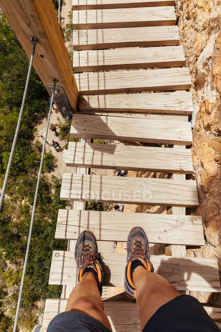 Top view of crop unrecognizable man legs standing on high mountain bridge over a river. — Stock Photo