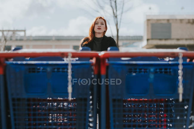 Atraente jovem mulher pensativa com cabelos vermelhos e carrinhos de compras no estacionamento em dia nublado — Fotografia de Stock