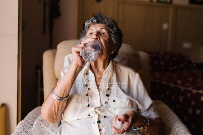 Mujer de pelo gris en camisa blanca bebiendo pastillas con agua de la botella, sentado en el sillón y mirando hacia otro lado en el apartamento - foto de stock