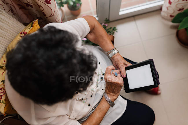Сверху пожилая седовласая женщина в рубашке сидит на кресле и читает электронную книгу в квартире — стоковое фото