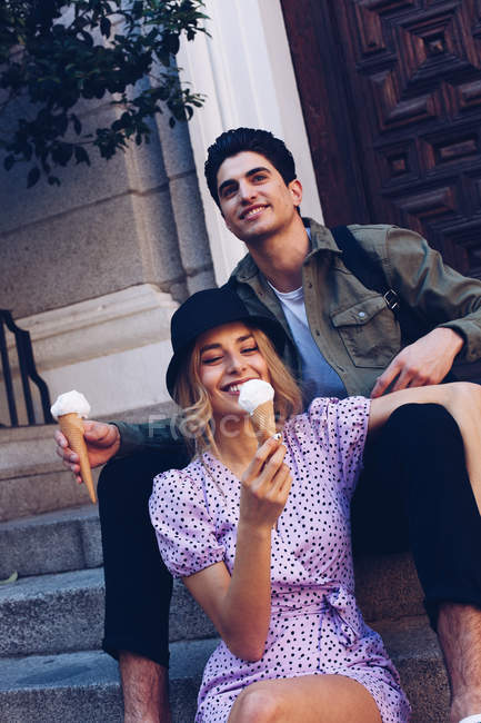 Веселая молодая привлекательная женщина и ее парень едят мороженое, сидя на улице — стоковое фото