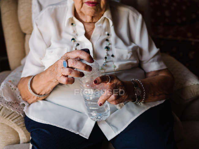 Cropped image of woman in white shirt drinking pills with water from bottle, sitting on armchair in apartment — Stock Photo