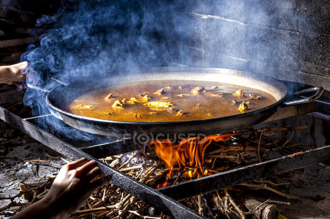 Colheita mãos irreconhecíveis segurando grande panela de ferro com caldo de ebulição para cozinhar paella sobre fogo aberto com madeira — Fotografia de Stock