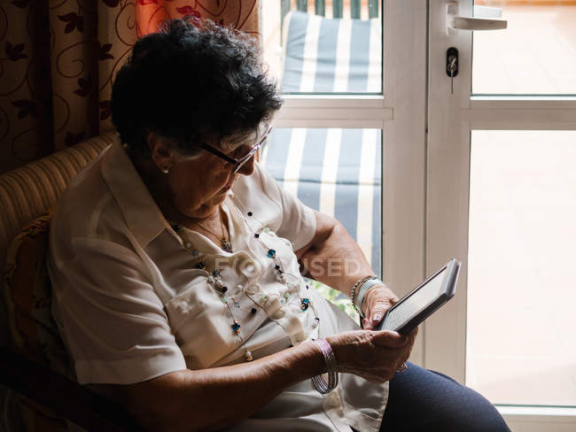 Anziana donna dai capelli grigi in camicia e occhiali seduta su poltrona e lettura e-book in appartamento — Foto stock