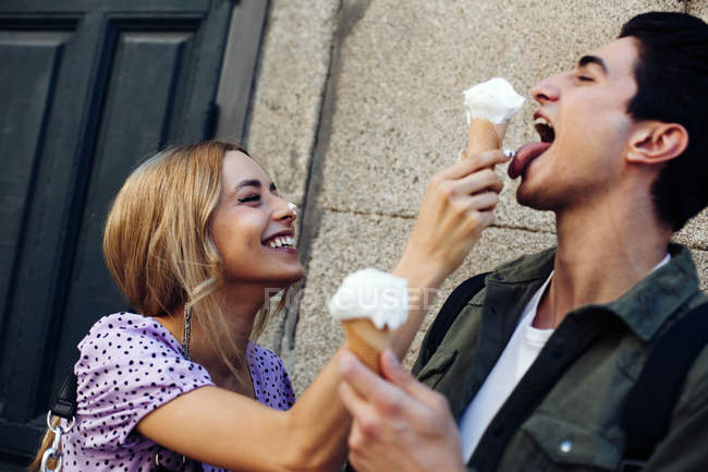 Vista lateral de alegre joven atractiva mujer alimentación novio por helado al aire libre - foto de stock