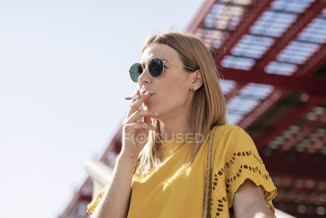 Молода стильна жінка в сонцезахисних окулярах курить сигарету на відкритому повітрі — стокове фото