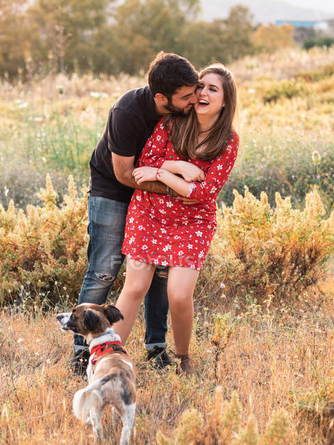 Jeune couple heureux étreignant et s'amusant avec petit chien parmi l'herbe haute dans la campagne — Photo de stock