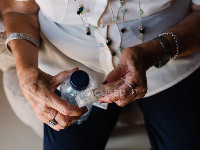 Cropped image of senior woman in white shirt holding pills and water from bottle, sitting on armchair in apartment — Stock Photo