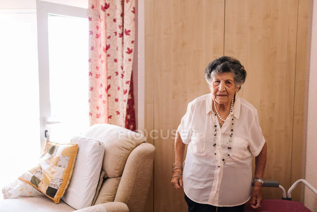 Mujer mayor en camisa blanca con la mano en la cintura de pie mientras se apoya en el sillón en casa y mirando a la cámara - foto de stock