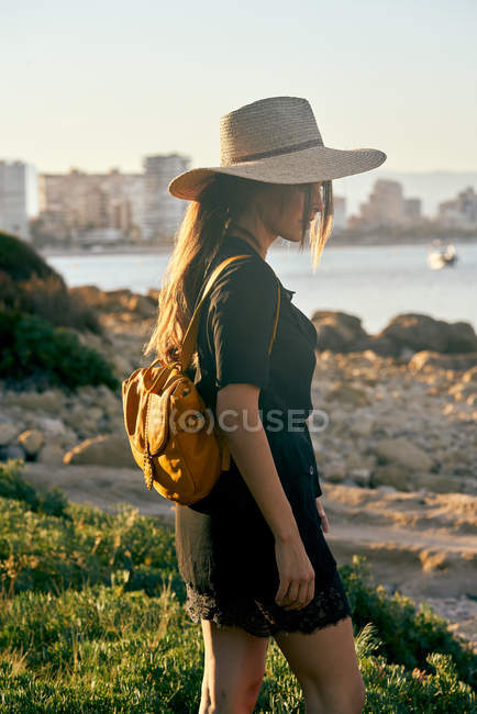 Turista feminino usando chapéu de palha e mochila de pé perto da praia — Fotografia de Stock